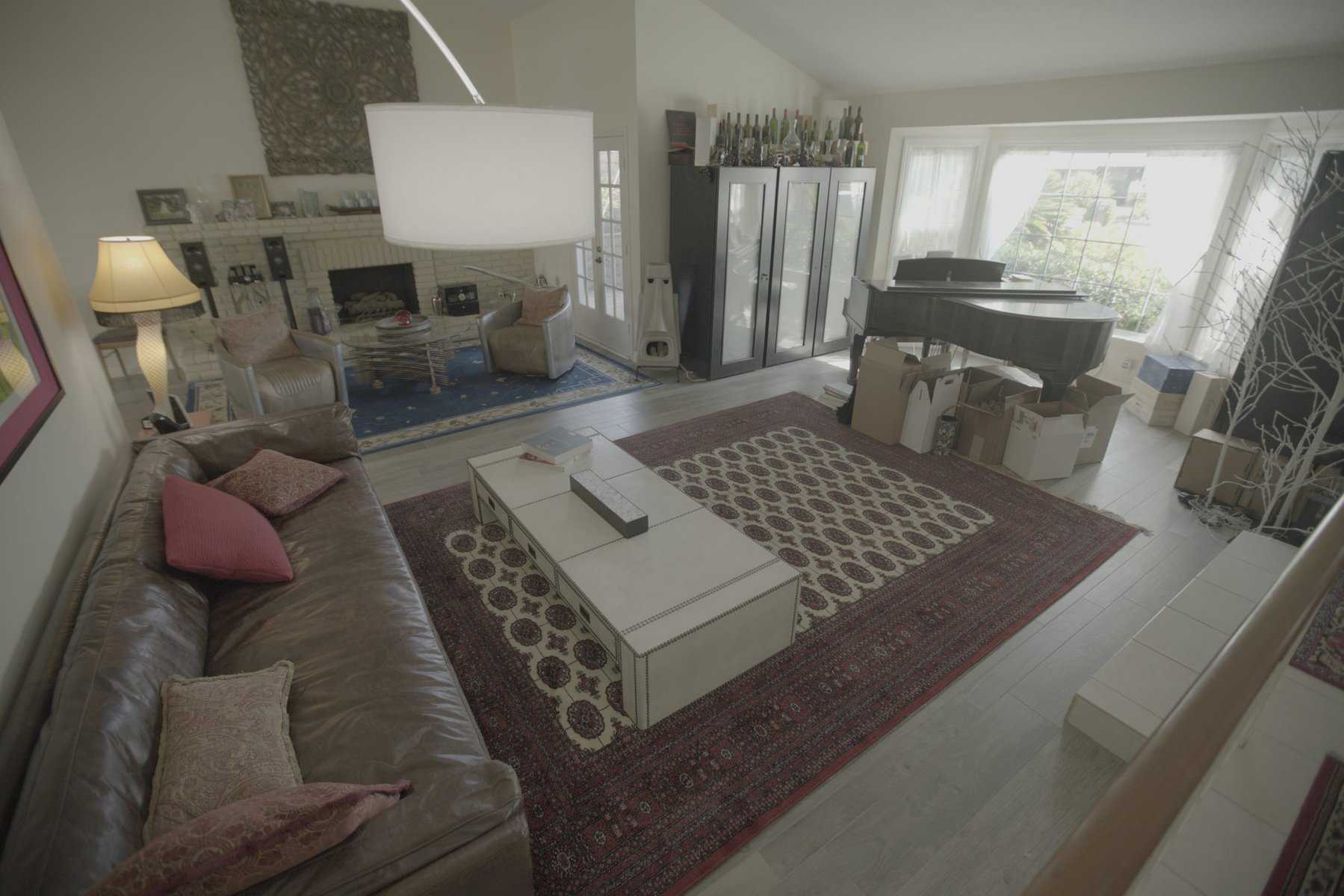 Cluttered living room with a leather sofa, patterned rugs, grand piano, and boxes near a large window