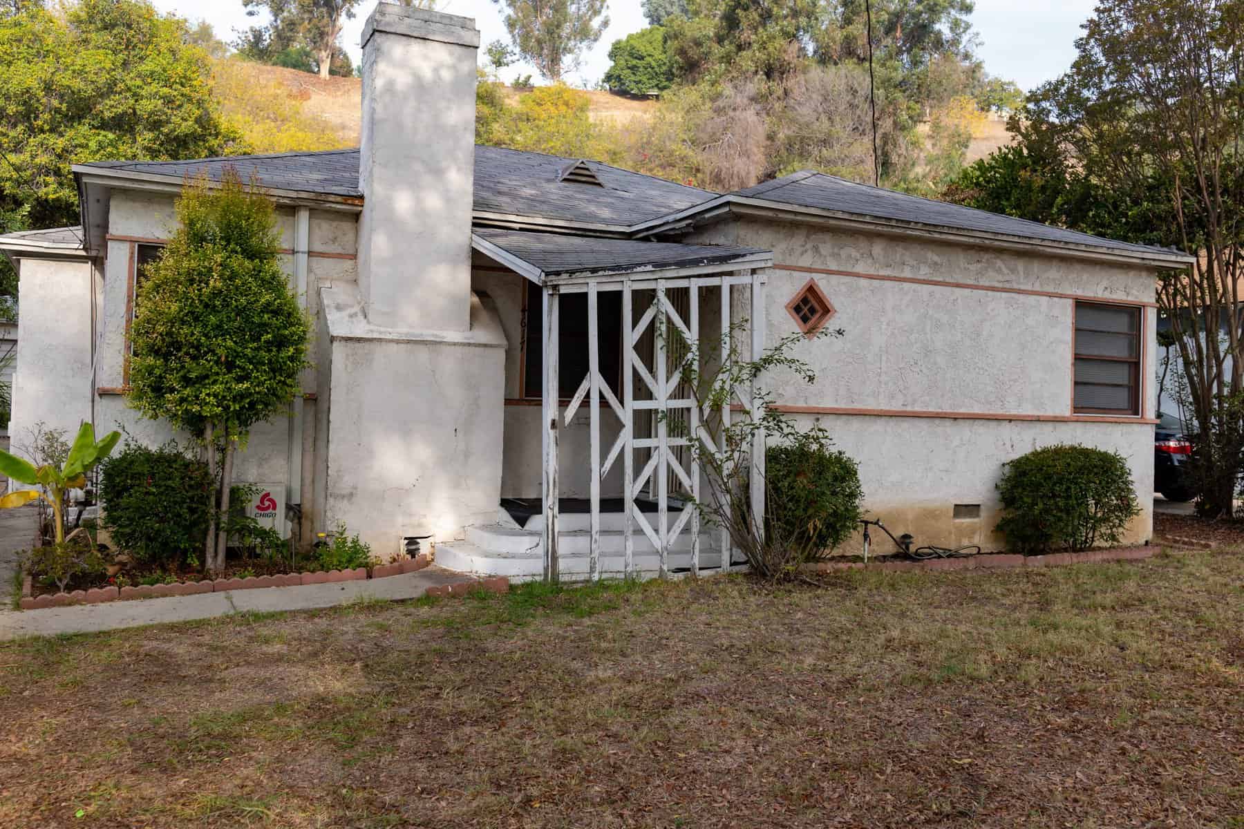 Aging single-story house with stucco exterior, chimney, and a small gated front porch